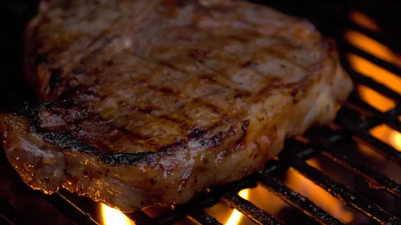 Thick bone-in T-Bone steak sizzling on a live-fire grill with glowing orange flames